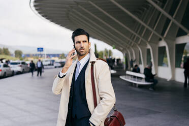 Busy man with dark hair in suit and coat speaking on phone while standing near airport terminal at daytime - ADSF19300