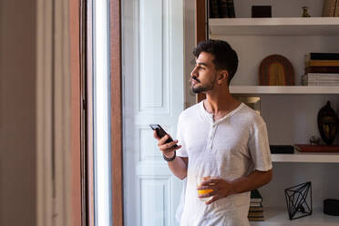 Junger bärtiger Mann mit einem Glas Fruchtsaft und einem Smartphone in der Hand, der am Fenster steht und interessiert in die Ferne blickt, wenn er morgens zu Hause ist - ADSF19306