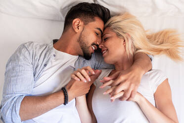 From above of cheerful couple in casual wear lying on soft bed at home and delicately cuddling with closed eyes - ADSF19312