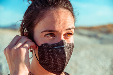 Side view of confident young sportswoman in black protective mask listening to music with wireless earbuds while recreating during training in rough desert badlands looking away - ADSF19324