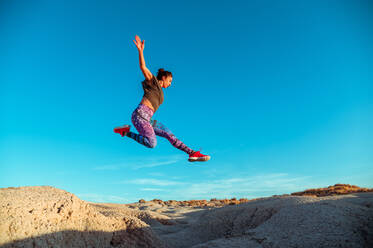 From below full body of active energetic female in sportswear leaping high above sandy slope of desert terrain against blue cloudless sky - ADSF19325