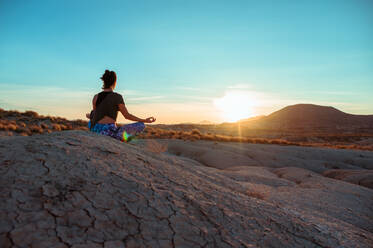 Rückenansicht einer nicht erkennbaren Frau in Sportkleidung, die in Lotus-Pose mit Mudra-Geste in einer rauen, hügeligen, steinigen Landschaft bei Sonnenuntergang meditiert - ADSF19326