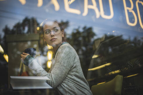 Blick durch Glas auf eine entspannte Frau in Freizeitkleidung, die mit einem aufgeschlagenen Buch in einem gemütlichen Café am Tisch sitzt und ein aromatisches Getränk genießt, während sie wegschaut - ADSF19328