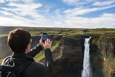 Tourist taking photo of Haifoss waterfall through mobile phone while standing at Iceland - NEKF00044