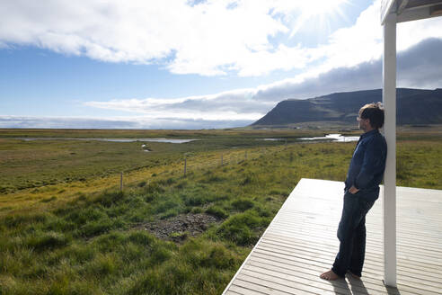 Älterer Mann betrachtet die Aussicht, während er sich auf einen Pfahl in den Westfjorden, Island, stützt - NEKF00060