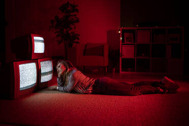 Young woman with hand in hair lying on floor in front of old TV's at living room - GIOF10172
