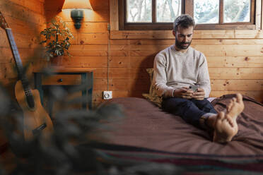 Junger Mann benutzt ein Mobiltelefon, während er in einer Holzhütte auf dem Bett sitzt - JPTF00620