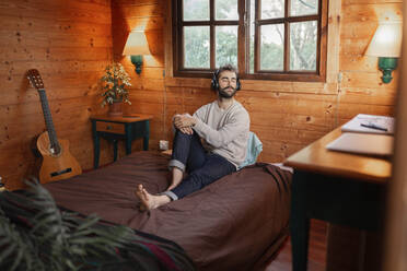 Junger Mann mit geschlossenen Augen Musik über Kopfhörer auf dem Bett in einer Holzhütte - JPTF00622
