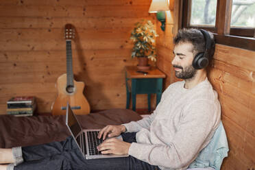 Lächelnder junger Mann, der einen Laptop benutzt und auf einem Bett in einer Blockhütte Musik hört - JPTF00623
