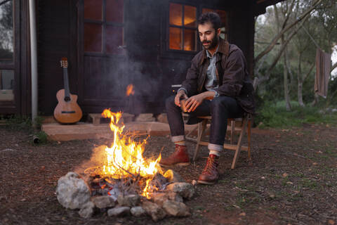 Young man with coffee cup sitting on chair near bonfire by wooden house stock photo