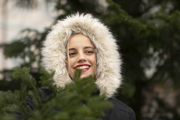 Smiling young woman in warm clothing day dreaming by tree during Christmas - SGF02720