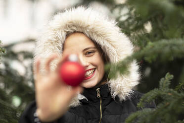 Lächelnde junge Frau in Pelzjacke hält Weihnachtsschmuck in der Nähe des Baumes - SGF02722