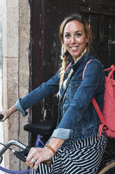 Smiling female tourist sitting on bicycle with backpack - JMPF00755