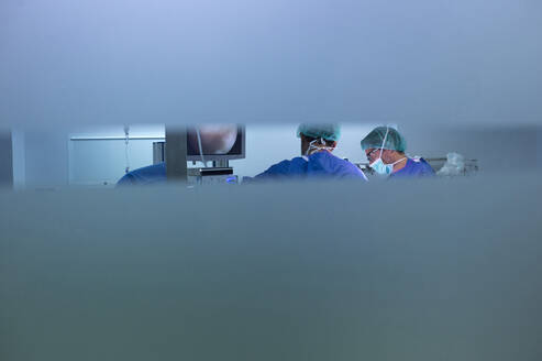 Männlicher Orthopäde, der durch ein Glasfenster bei einer Operation in der Notaufnahme gesehen wird - JCMF01710
