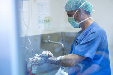 Male orthopedic surgeon washing hand with soap by sink in hospital - JCMF01716