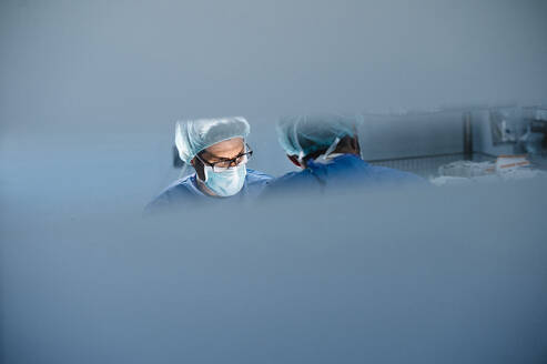 Male surgeons seen from glass while performing operation in hospital - JCMF01726