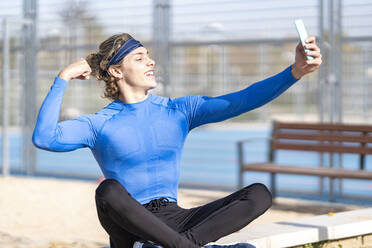 Lächelnder männlicher Sportler zeigt seinen Bizeps, während er ein Selfie im öffentlichen Park an einem sonnigen Tag macht - GGGF00517