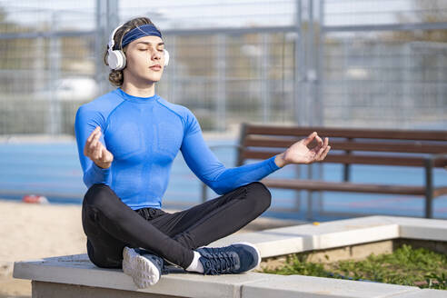 Young sportsman listening music while sitting in lotus position on retaining wall during sunny day - GGGF00518