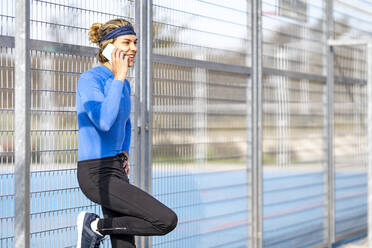Lächelnder männlicher Sportler beim Telefonieren am Zaun an einem sonnigen Tag - GGGF00523