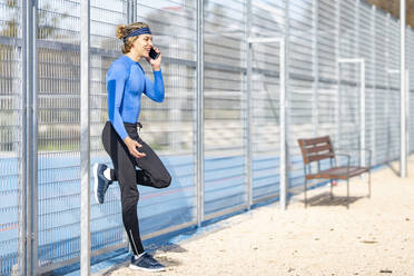 Lächelnder männlicher Sportler, der mit seinem Handy telefoniert, während er auf einem Bein am Zaun steht, an einem sonnigen Tag - GGGF00524