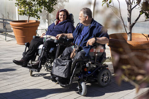 Disabled woman and man holding hands while sitting on wheelchair during sunny day stock photo