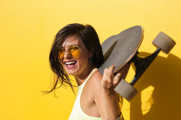 Porträt einer jungen schönen Frau, die mit einem Longboard in den Händen an einer gelben Wand steht - AODF00049