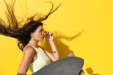 Porträt einer jungen schönen Frau, die sich vor einer gelben Wand mit einem Longboard in der Hand die Haare wirft - AODF00050