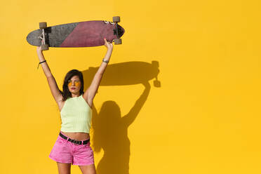 Porträt einer jungen Frau, die vor einer gelben Wand steht und ein Longboard in den Händen hält - AODF00051