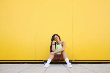Porträt eines schönen Mädchens, das allein auf einem Longboard vor einer gelben Wand sitzt - AODF00052