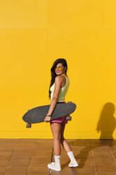 Porträt einer jungen Frau, die vor einer gelben Wand steht und ein Longboard in den Händen hält - AODF00054