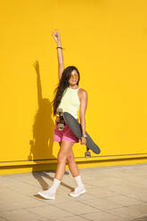 Portrait of young woman walking along yellow wall with longboard in hand - AODF00061