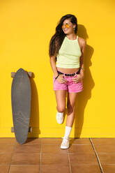 Junge schöne Frau posiert mit Longboard vor einer gelben Wand - AODF00066