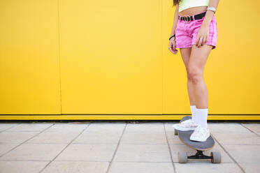 Low section of young woman standing on longboard in front of yellow wall - AODF00071