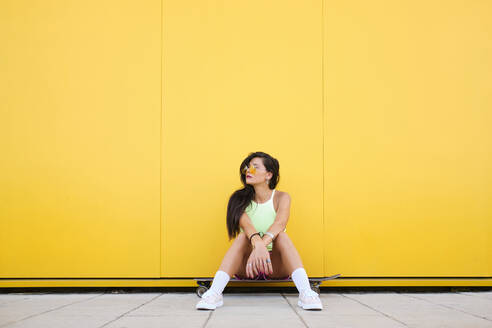 Porträt eines schönen Mädchens, das allein auf einem Longboard vor einer gelben Wand sitzt - AODF00072
