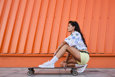 Portrait of beautiful girl crouching beside longboard in front of corrugated iron wall - AODF00080