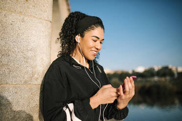 Lächelnde Sportlerin mit Handy, die Musik hört, während sie sich an eine Säule lehnt, an einem sonnigen Tag - GRCF00540