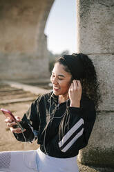 Glückliche Sportlerin, die an einem sonnigen Tag In-Ear-Kopfhörer einstellt, während sie ein Mobiltelefon benutzt - GRCF00542