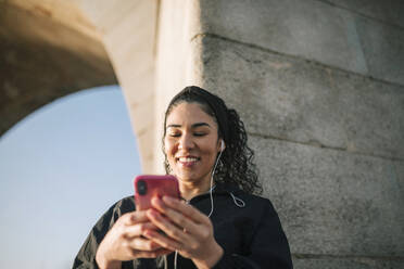 Lächelnde Sportlerin, die ein Mobiltelefon benutzt, während sie an der Wand Musik hört - GRCF00543