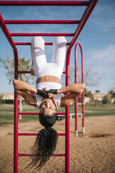 Sportlerin hängt kopfüber an einem Klettergerüst in einem öffentlichen Park an einem sonnigen Tag - GRCF00555