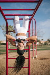 Sportswoman hanging upside down on monkey bars in public park during sunny day - GRCF00555