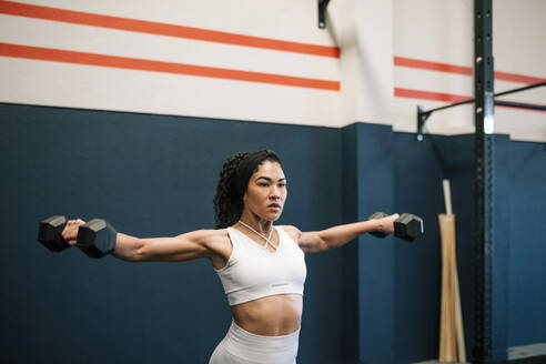 Female athlete working out with dumbbells against wall in gym - GRCF00563