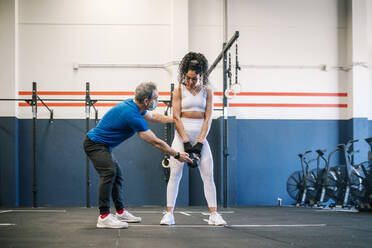 Male fitness in mask instructor explaining sportswoman about kettlebell in gym - GRCF00578