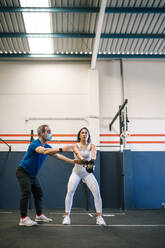 Männlicher Fitnesstrainer trainiert eine Sportlerin mit einer Kettlebell im Fitnessstudio während COVID-19 - GRCF00580