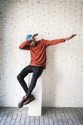 Man dabbing while sitting on pedestal against white brick wall - RCPF00487