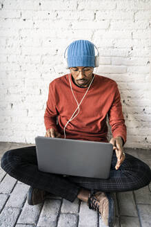 Freelance worker using laptop while listening music through headphones against brick wall - RCPF00492