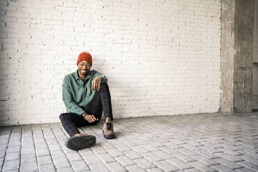 Happy man in knit hat sitting on floor against white brick wall - RCPF00498
