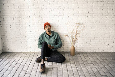 Mid adult man with blank expression sitting by dried plant vase against white brick wall - RCPF00502