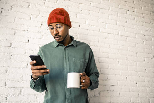 Mann mit Kaffeetasse und Smartphone vor weißer Backsteinwand - RCPF00503
