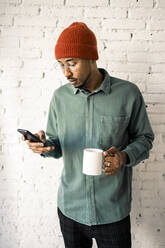 Mittlerer erwachsener Mann, der einen Kaffee trinkt, während er ein Mobiltelefon vor einer weißen Backsteinmauer benutzt - RCPF00505