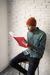 Man wearing eyeglasses reading book against white brick wall - RCPF00506